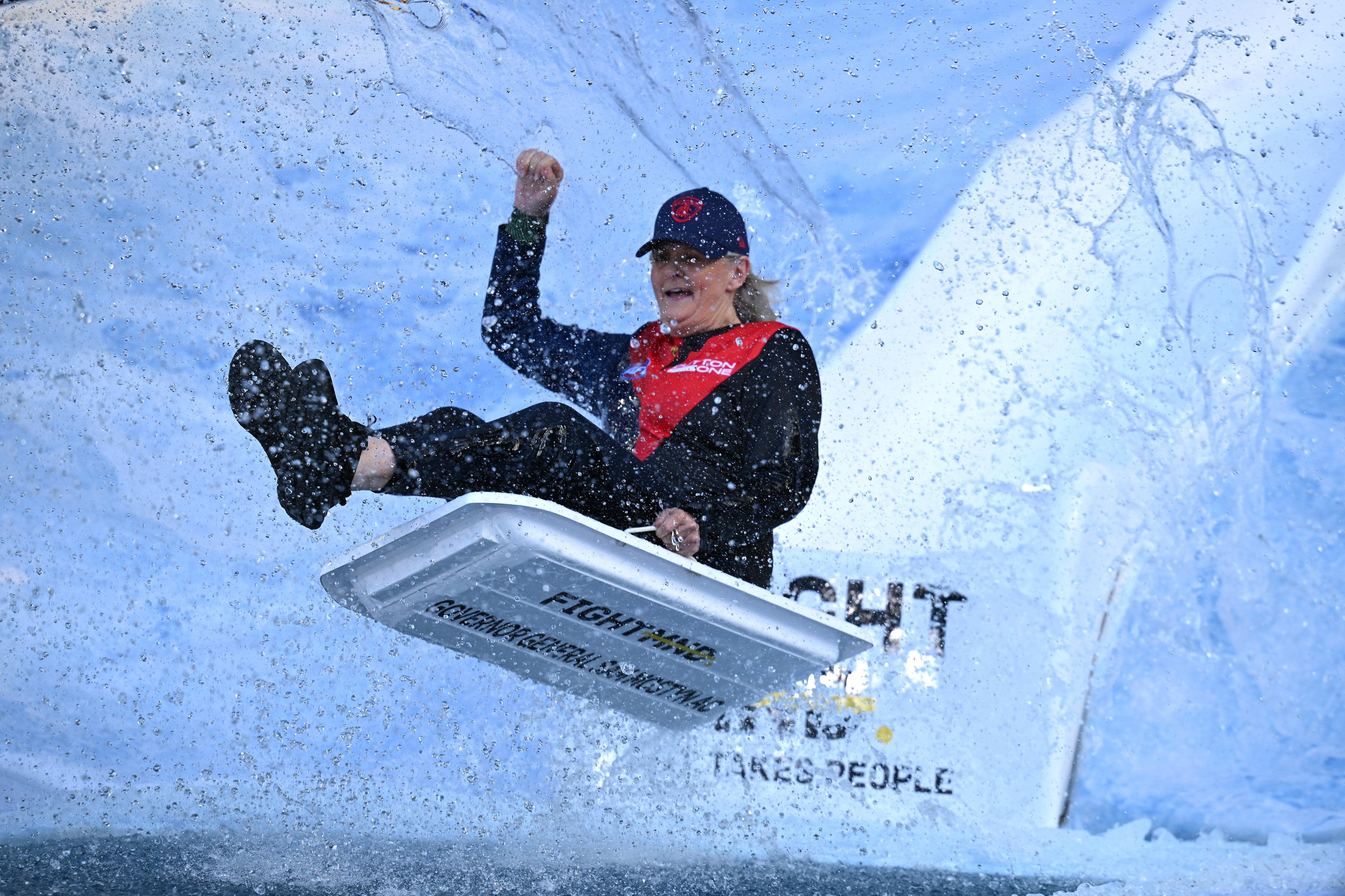A blonde woman in a navy cap, blue and red top and blue pants sits on a white sled with her fist in the air amid water droplets.