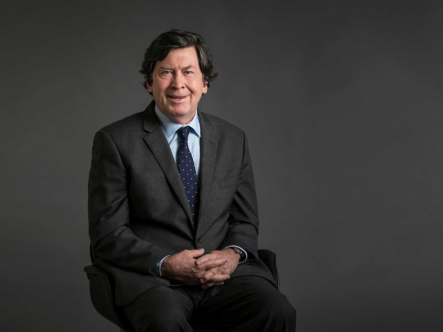 A man with dark brown hair wearing a grey suit and blue shirt and tie sitting on a seat in front of a grey background