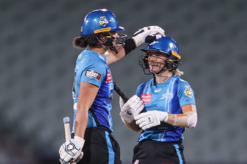 A woman in a blue cricket uniform pats her teammate on the helmet.
