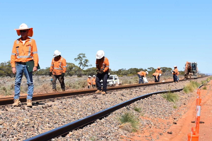 A group of men in high-vis shirts standing on a rail road. There is red dust around them.