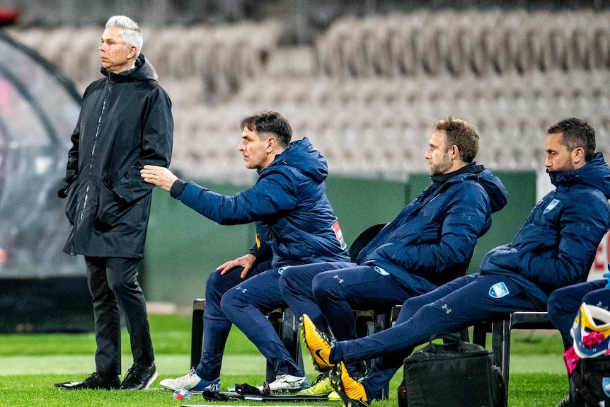 Steve Corica stands wearing a jacket next to three men who are sitting on chairs