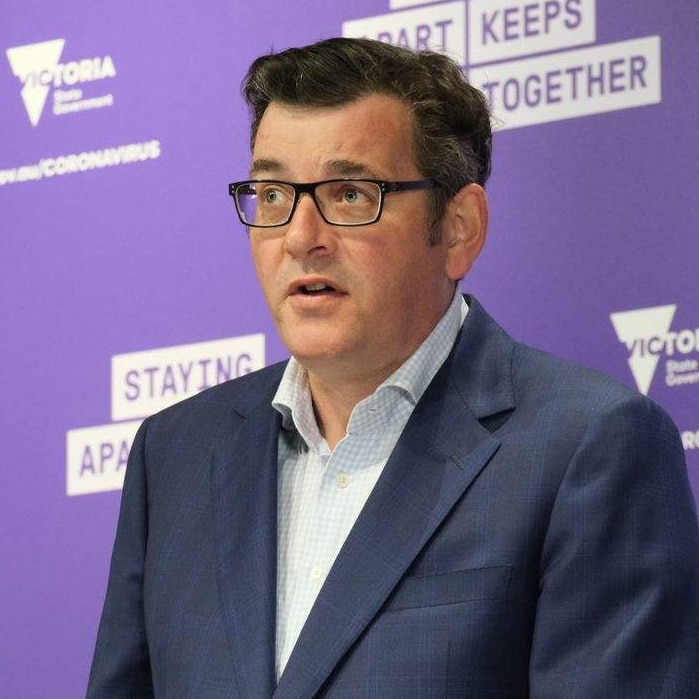 Daniel Andrews, dressed in a shirt and jacket, stands at a lectern with microphones in front of a purple background.