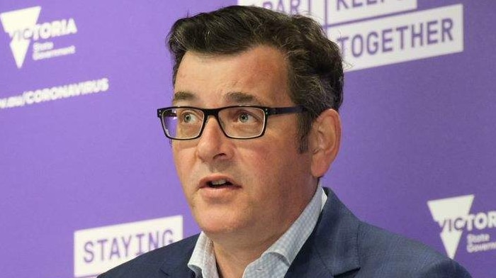 Daniel Andrews, dressed in a shirt and jacket, stands at a lectern with microphones in front of a purple background.