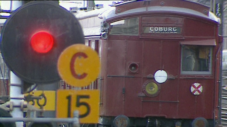 A burgundy red train on tracks with 'Coburg' sign, red train stoplight and assorted yellow signs in foreground.