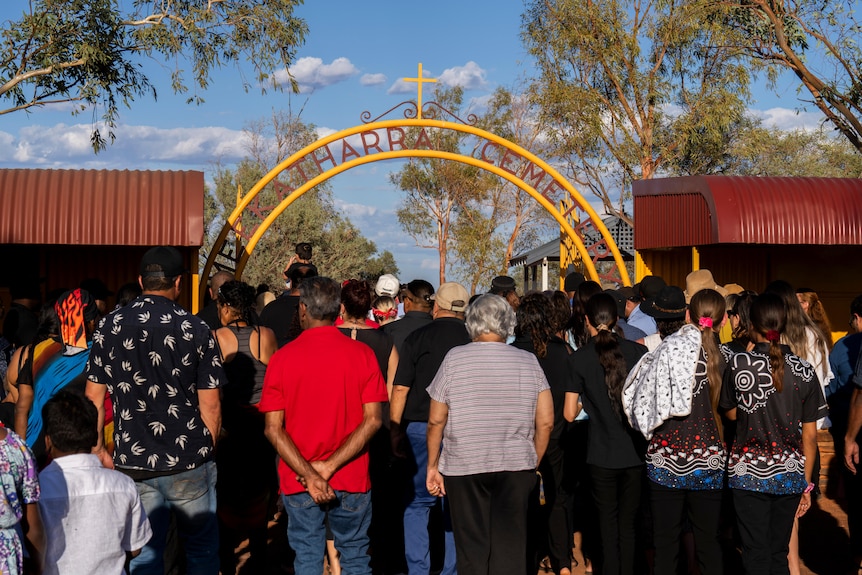 A crowd of people with their backs to the camera in front of a cemetery entrance.