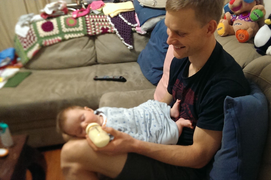 A man in a black t-shirt smiling warmly as he bottle-feeds a baby girl.