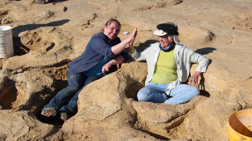 Two women sitting in huge dinosaur footprints high five each other.