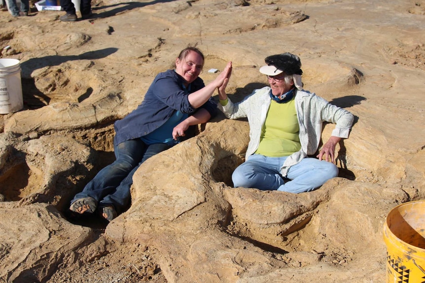 Two women sitting in huge dinosaur footprints high five each other.