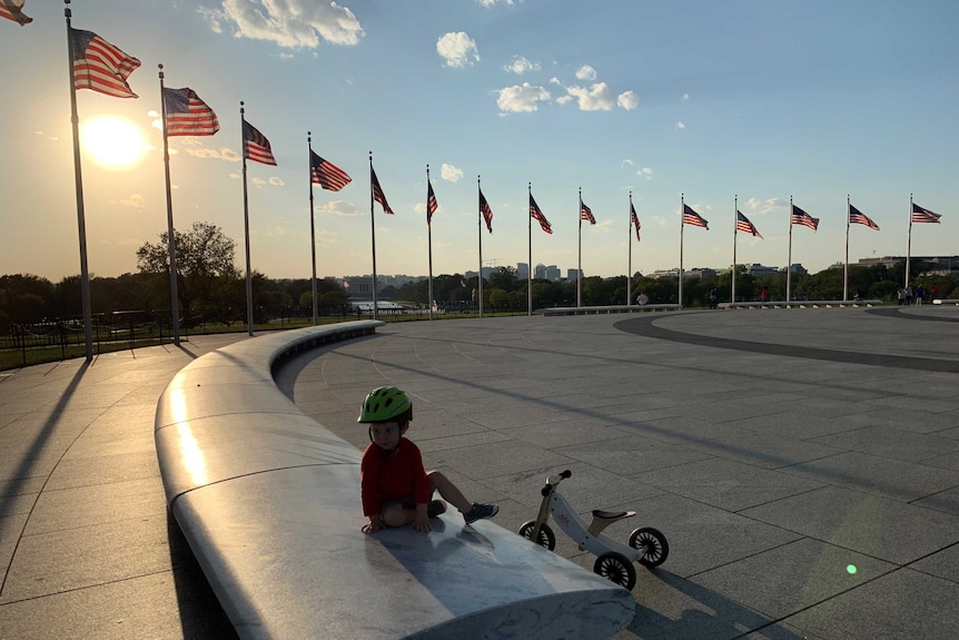 Little boy with bike and wearing bike helmet sitting on bench surrounded by American flags.