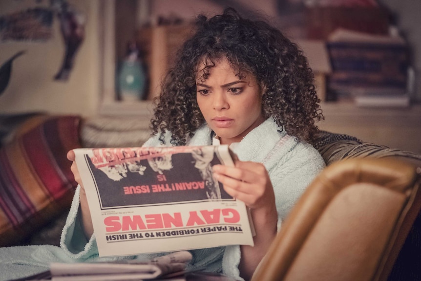 A still from the miniseries It's a Sin with a young woman reading a newspaper called "Gay News" and looking concerned