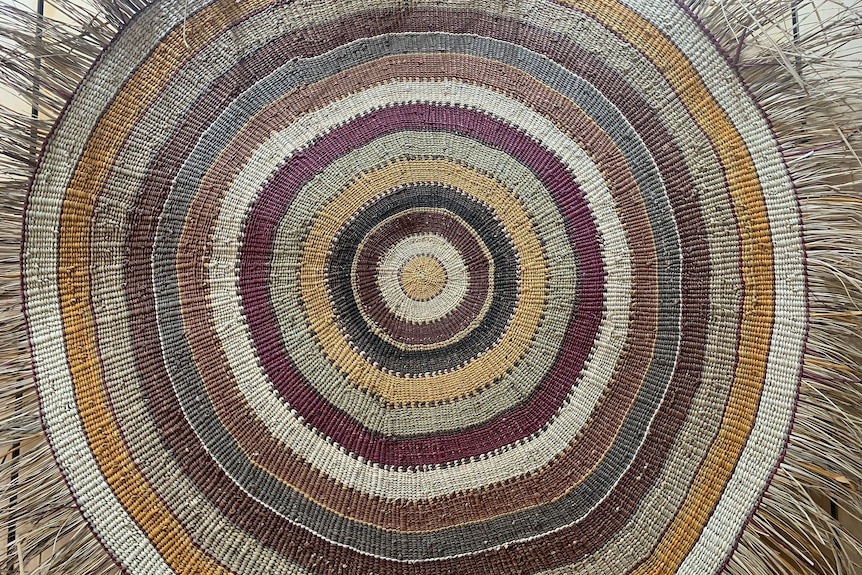 A woven piece of art hangs in an art centre. It is circular, and has green, yellow and red tones.