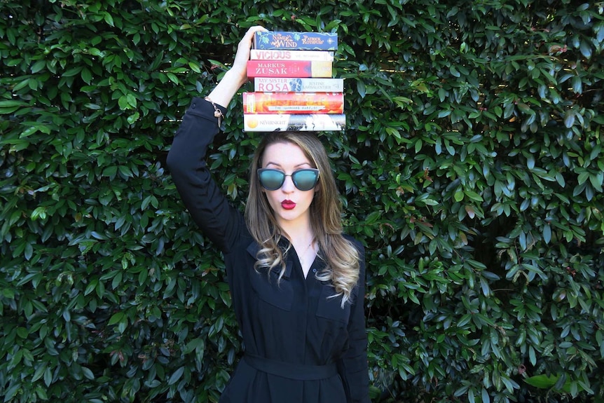 A young white woman with big sunglasses and a stack of books on her head, stands in front of a wall of bushes