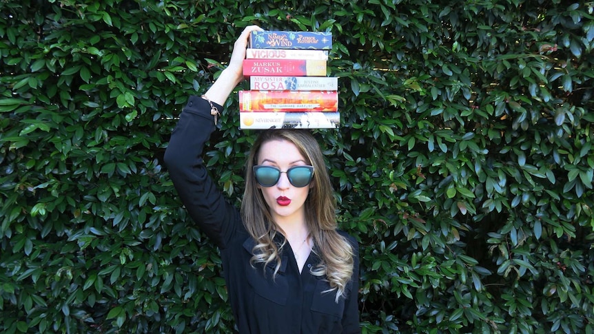 A young white woman with big sunglasses and a stack of books on her head, stands in front of a wall of bushes