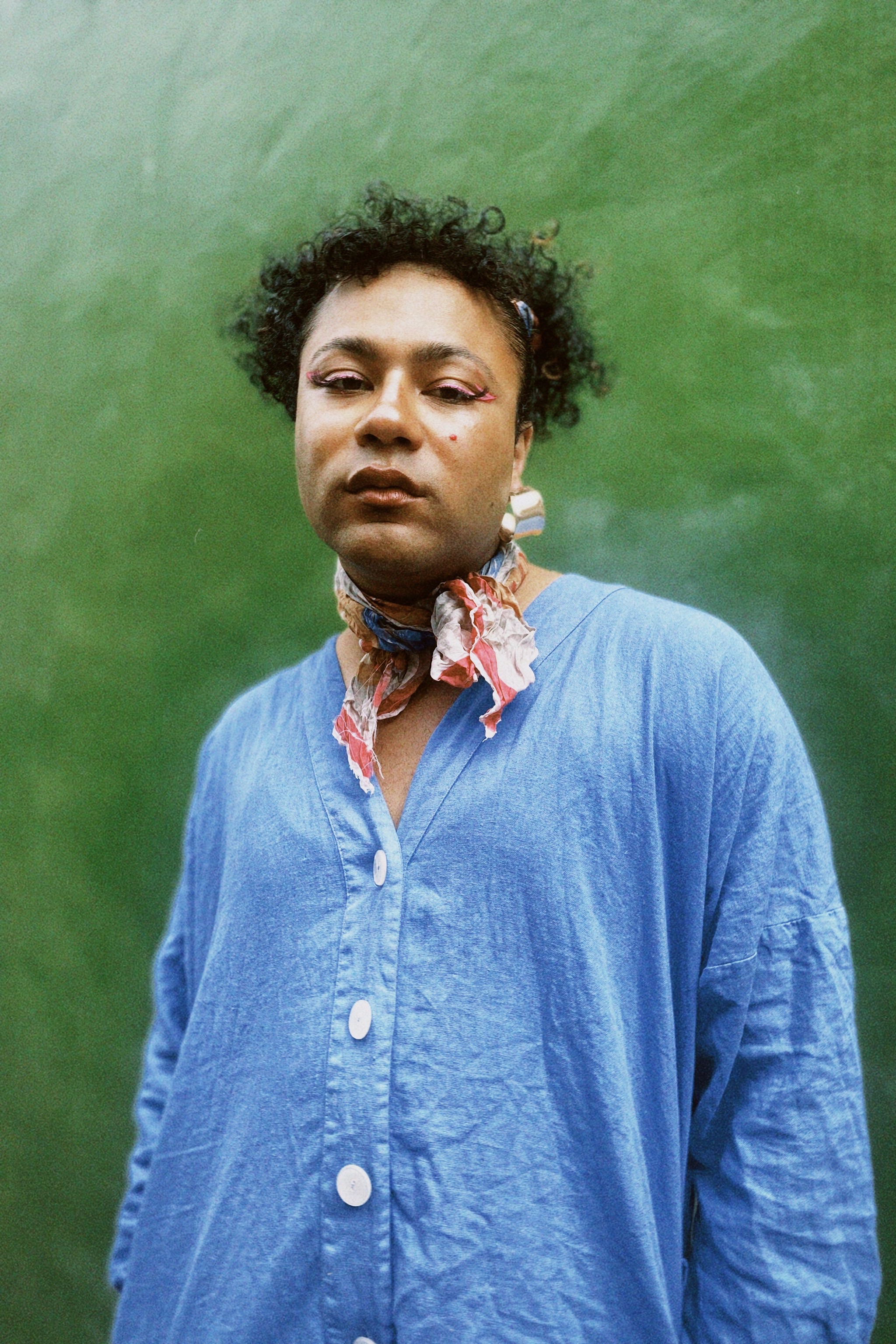 Trans woman with dark skin and curly hair wears colourful neck tie and blue shirt against a frosted green backdrop.