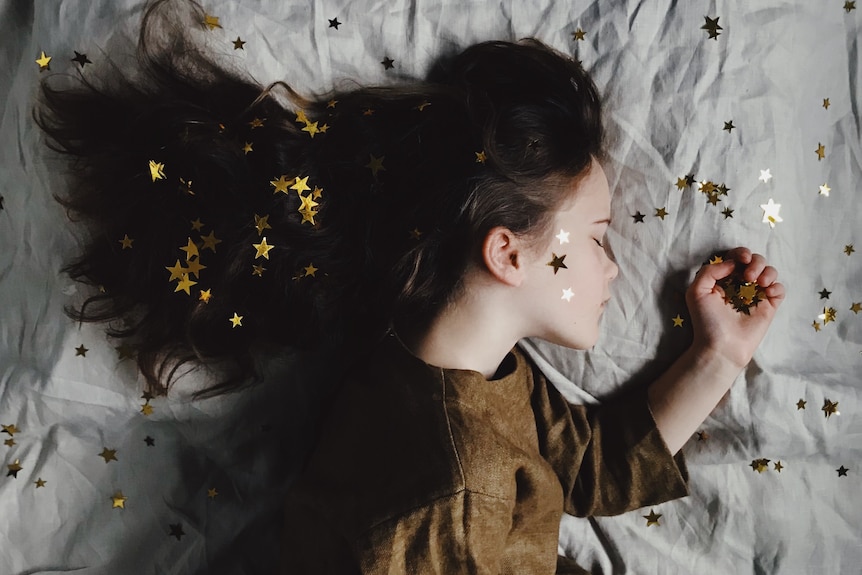 A photo of a young girl, shot from above, sleeping in bed, surrounded by gold star confetti