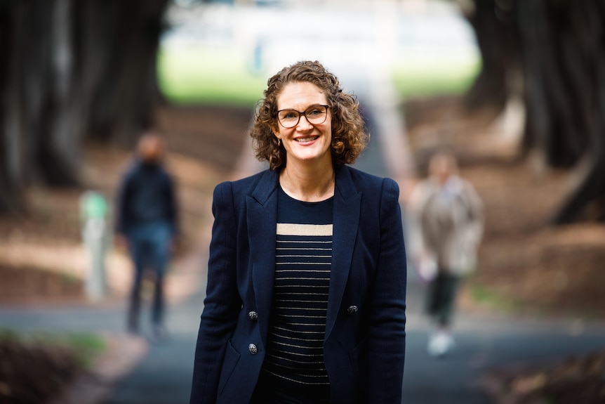 A woman with short curly hair, wearing dark framed spectacles and a navy blazer, walks along a tree lined path