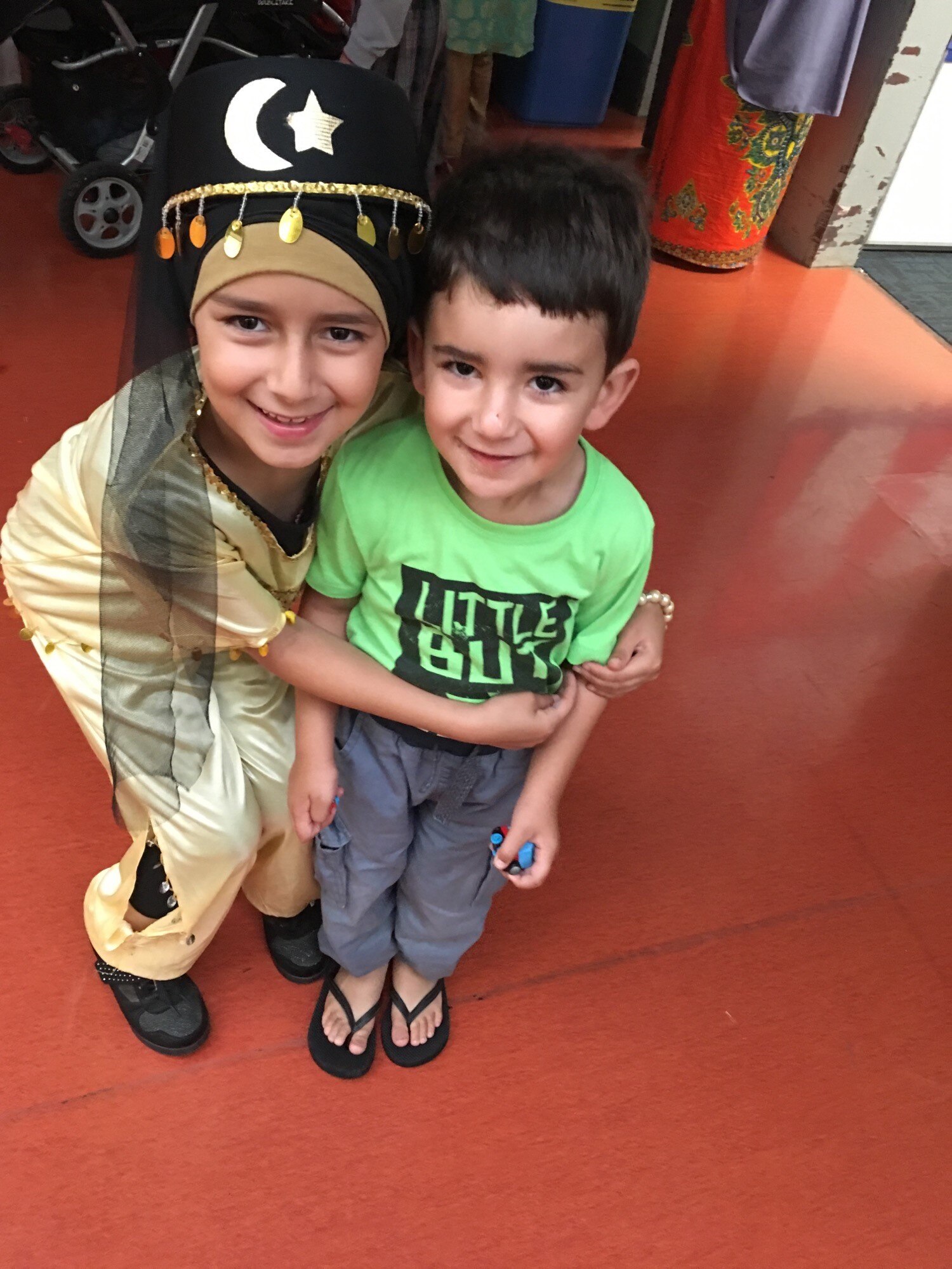 A young girl hugs her brother. They both smile. She wears a black hat with gold sequins. He wears a bright green t-shirt.