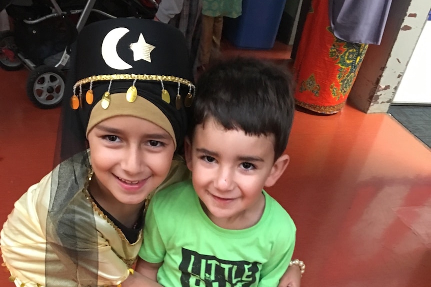 A young girl hugs her brother. They both smile. She wears a black hat with gold sequins. He wears a bright green t-shirt.