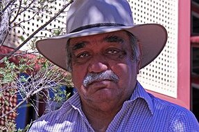 An Indigenous man looks into a camera, he is wearing a white broad-rimmed hat, and a stripy blue shirt.