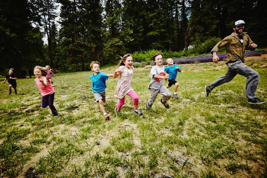 A group of young children and a man chase each other in a grassy field.