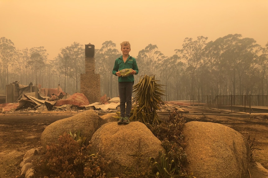 Boy holds football while standing on rock surrounded by bushfire damage, in story about children and bushfire trauma.