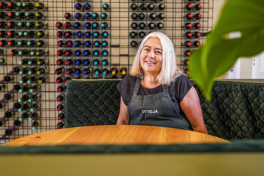 A woman with a blonde bob haircut wearing an apron sits in a velvet green restaurant booth with a wine rack behind her.