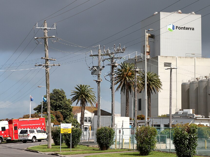 A large concrete building with Fonterra sign and cylinder vats on a street with cars in the driveway.