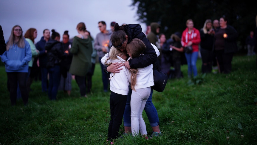 A woman stands on a grassy hill, embracing two children during a vigil for the victims of the Keyham mass shooting