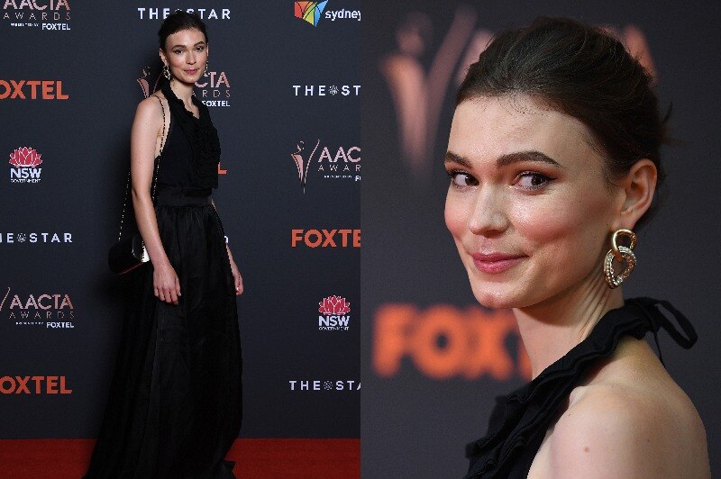 A woman in a formal black dress with gold earrings poses on the red carpet.