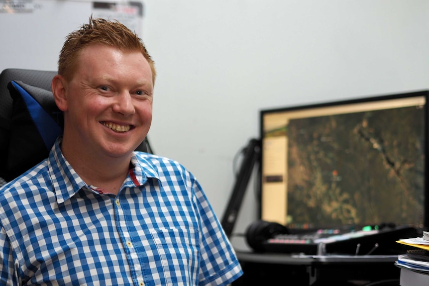 A man in a blue checked shirt sits in front of a computer and smiles at the camera