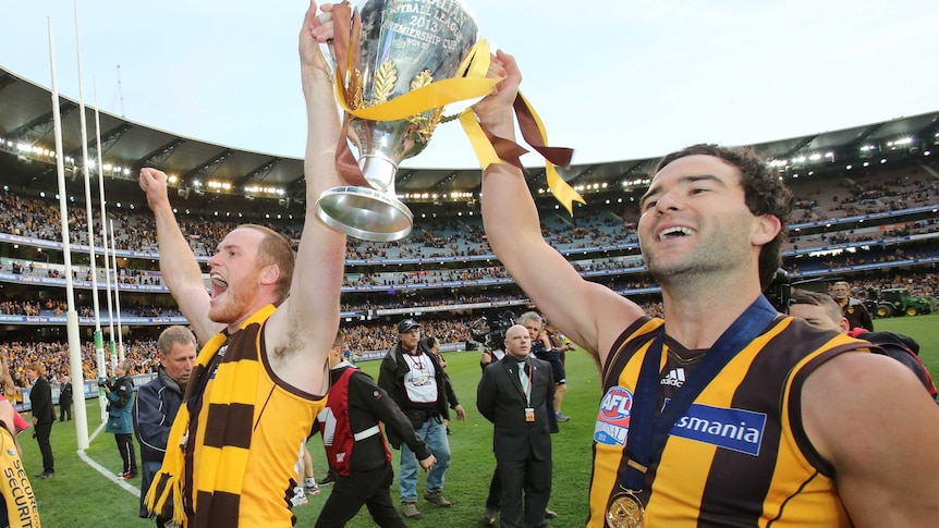Jarryd Roughead and Jordan Lewis face the crowd while lofting the premiership cup. Both are smiling.