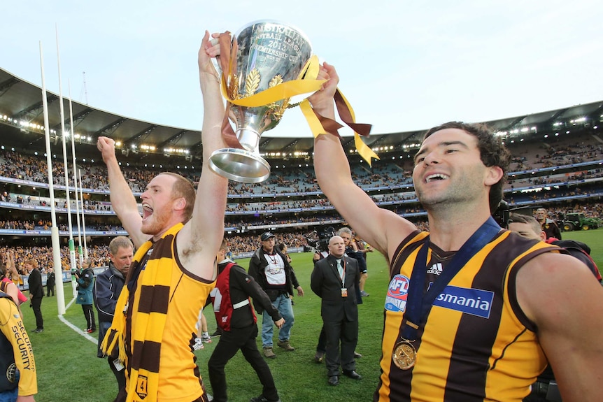 Jarryd Roughead and Jordan Lewis face the crowd while lofting the premiership cup. Both are smiling.