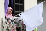 A woman wearing a headscarf stands with a white flag out the window of her apartment