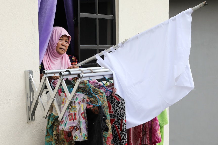 A woman wearing a headscarf stands with a white flag out the window of her apartment