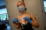 A person wearing a face mask and gloves stands in front of a mirror and adjusts a central line on their body. 