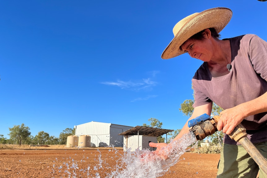 A woman wearing a wide-brimmed straw hat and T-shirt sprays water on her hand with a large hose, on a rural property.