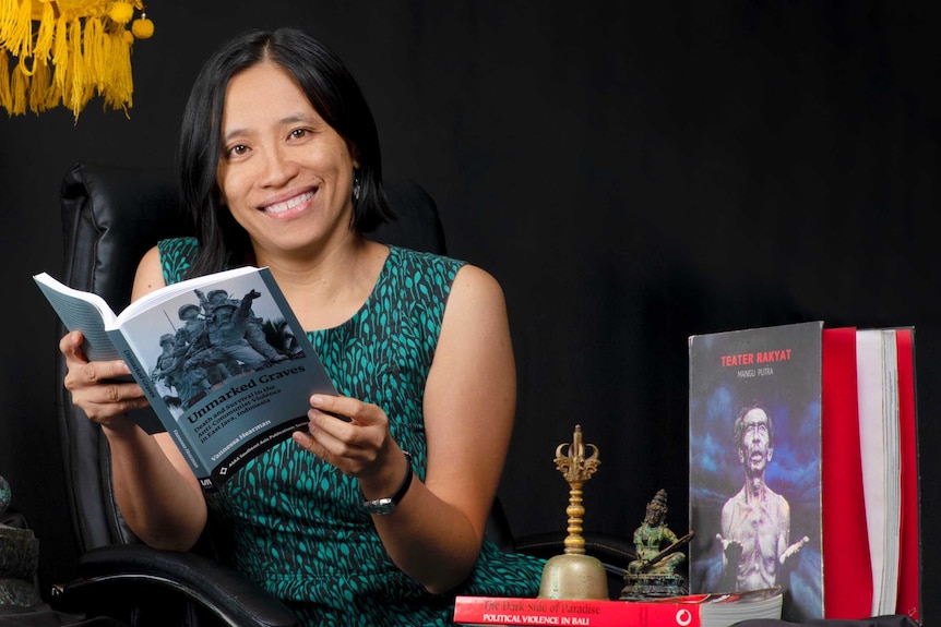 You view a woman holding a book and looking into the camera as she is surrounded by traditional Indonesian objects.
