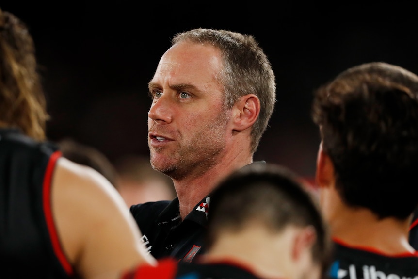A serious-looking AFL coach talks to a player at three quarter-time in a match as other players watch stand near him.