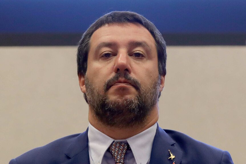 Matteo Salvini stands against a plain backdrop, and looks at the camera with serious expression.