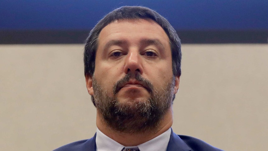 Matteo Salvini stands against a plain backdrop, and looks at the camera with serious expression.