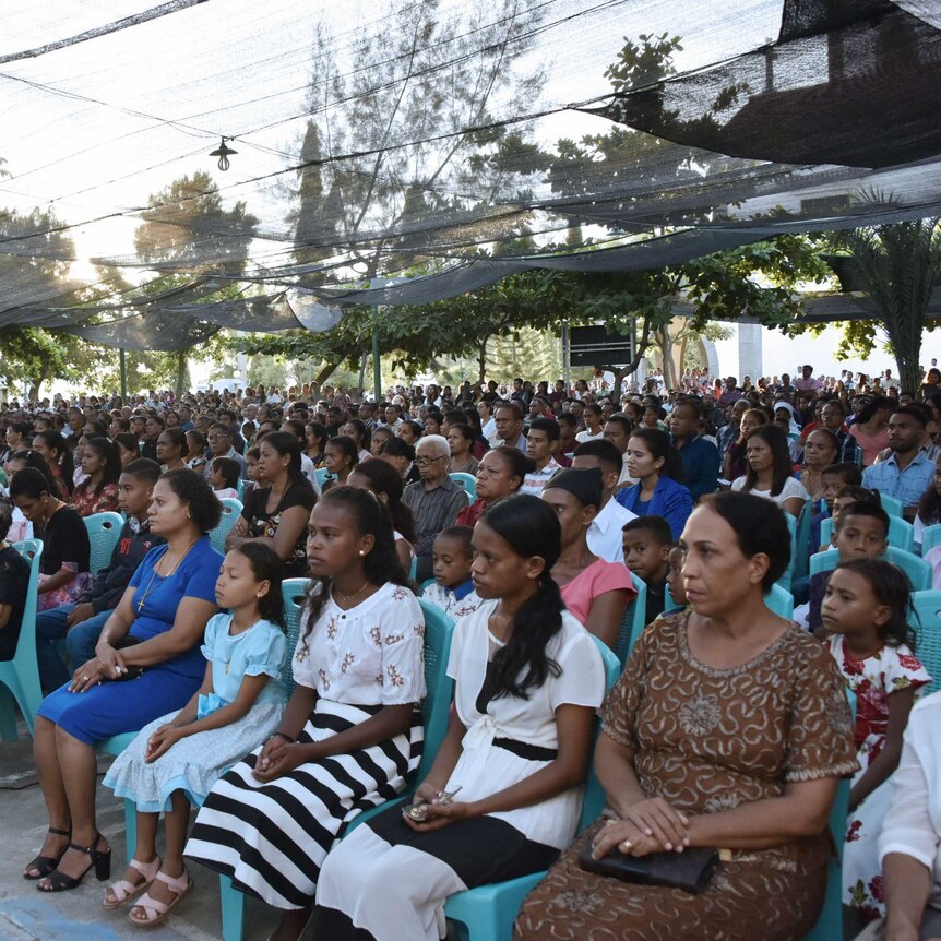 rows of seated people, mostly women and girls, dressed formally, in a town square