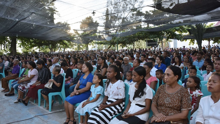 rows of seated people, mostly women and girls, dressed formally, in a town square