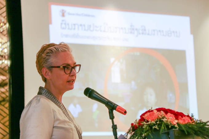 A woman with glasses speaks into a microphone in front of a slideshow