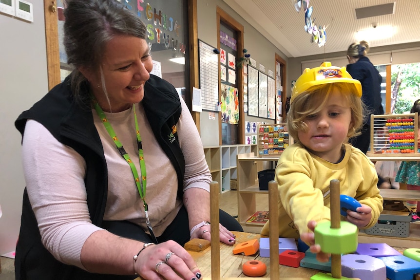 An smiling woman wearing a pink top, black jacket, green lanyard, plays with a toddler, colourful toys.