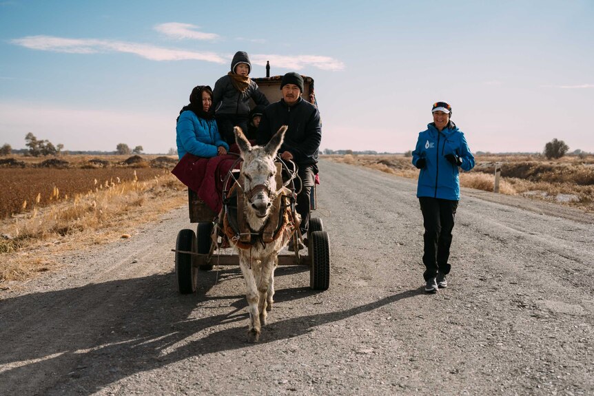 On an rural road, Mina Guli runs alongside three village locals who are sitting on a cart pulled by a donkey
