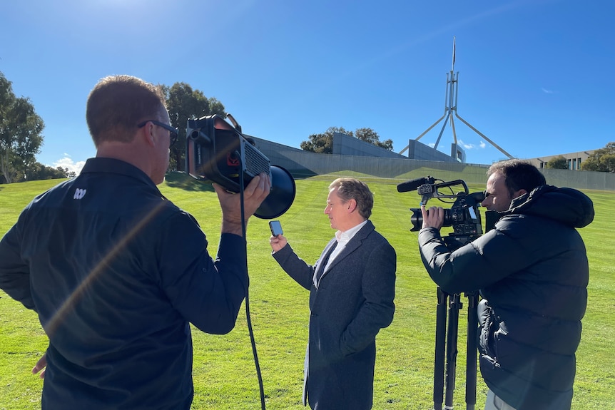 Two men holding cameras film man holding a mobile phone with Parliament House in the background.