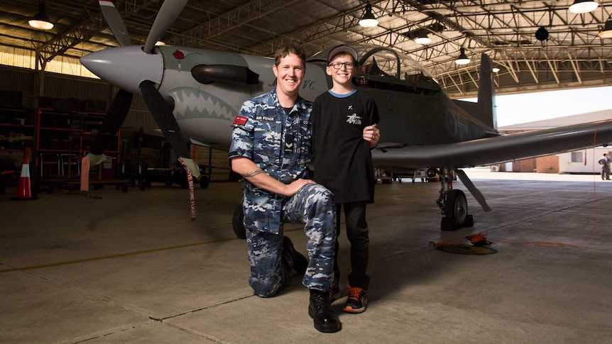 Trevor Connell kneels next to Finn Coker in front of a plane in a hangar.
