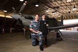 Trevor Connell kneels next to Finn Coker in front of a plane in a hangar.