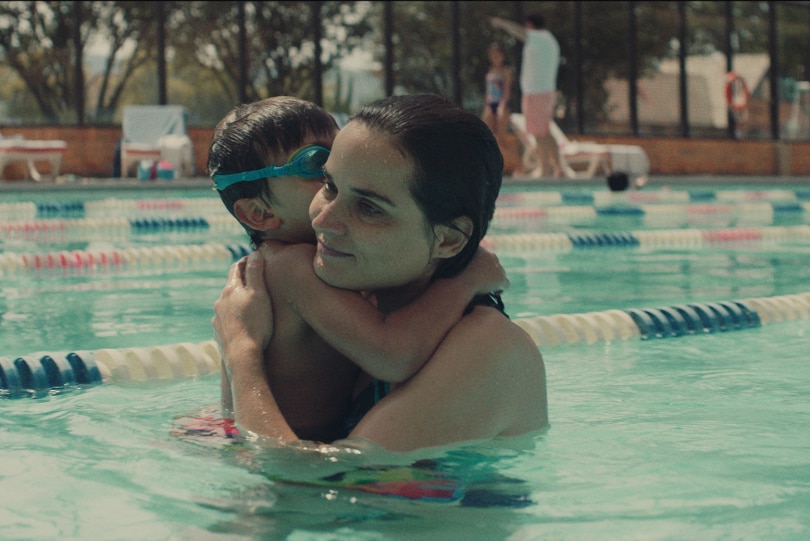 A woman with wet pulled back hair stands in lane of swimming pool carrying young boy wearing goggles on sunny day.