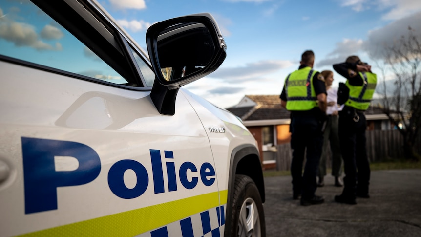 Two blurred police officers talk to a woman behind a white police car with blue writing